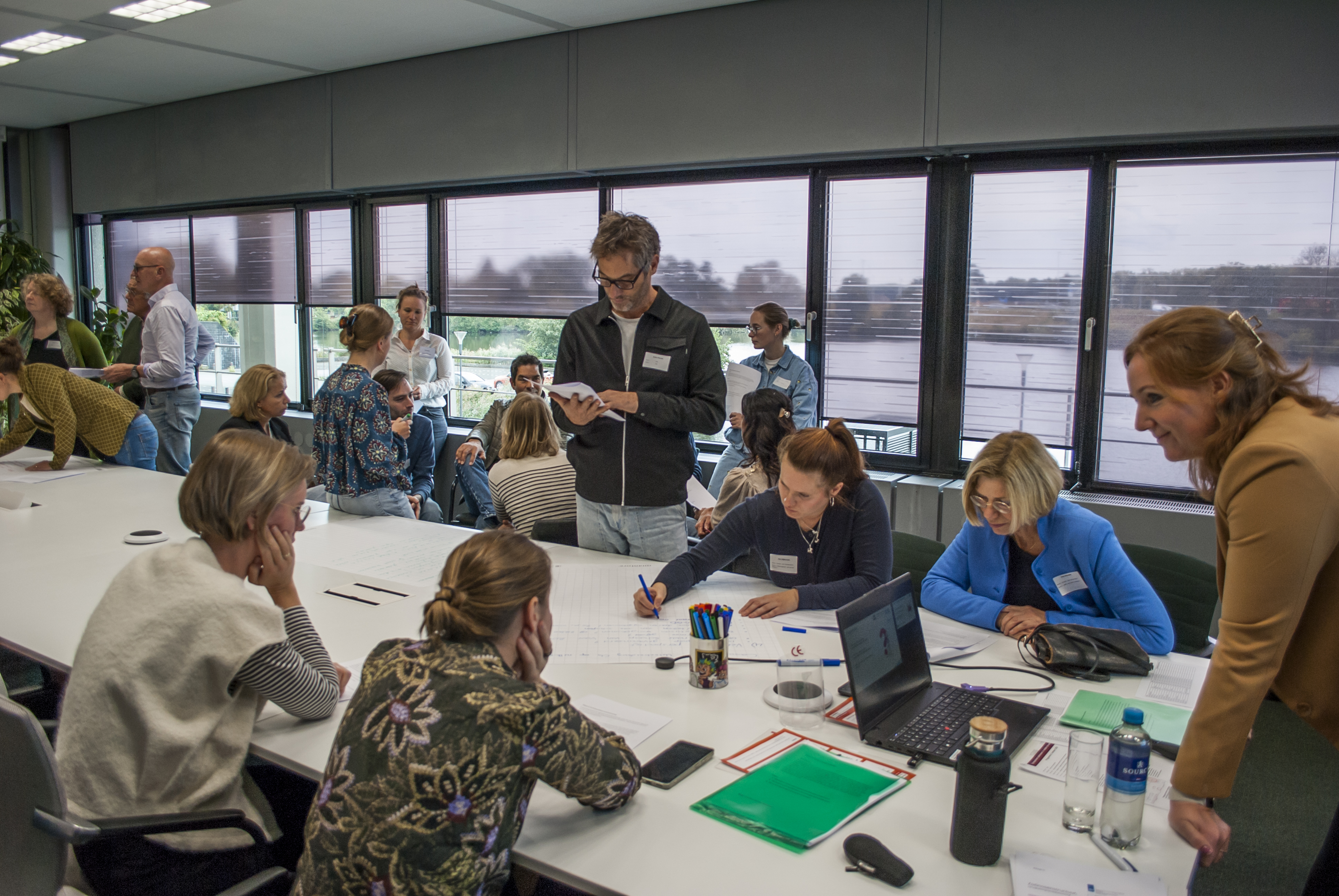 Mensen zijn om een tafel interactief bezig met een opdracht. Er is veel beweging in de zaal, sommige mensen zitten, andere staan.