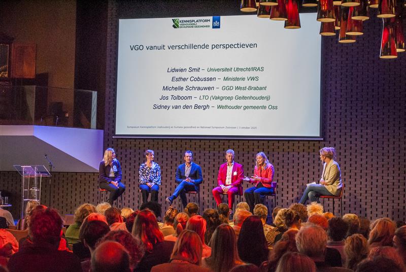 Er zitten 5 mensen op hoge krukken op een podium naast elkaar, zij zijn in gesprek met de dagvoorzitter