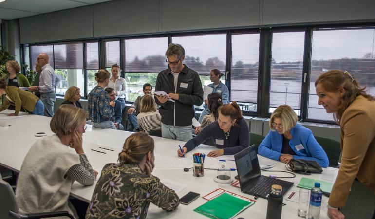 Mensen zijn om een tafel interactief bezig met een opdracht. Er is veel beweging in de zaal, sommige mensen zitten, andere staan.