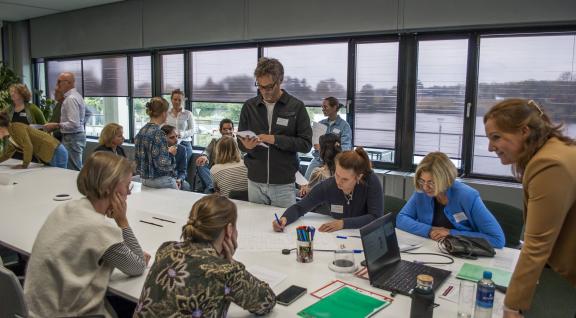 Mensen zijn om een tafel interactief bezig met een opdracht. Er is veel beweging in de zaal, sommige mensen zitten, andere staan.