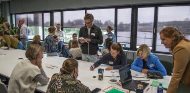 Mensen zijn om een tafel interactief bezig met een opdracht. Er is veel beweging in de zaal, sommige mensen zitten, andere staan.